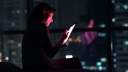 Young woman browsing web on tablet in bed at night with city lights