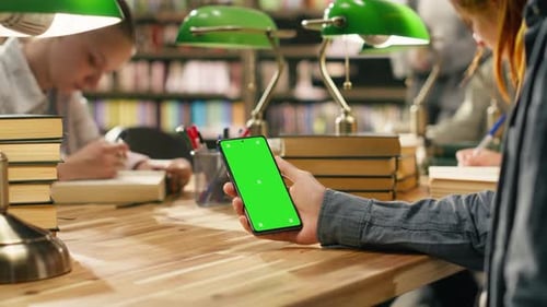 Vertikaler isolierter grüner Bildschirm auf einem Smartphone, das über dem hölzernen Bibliothekstisch gehalten wird