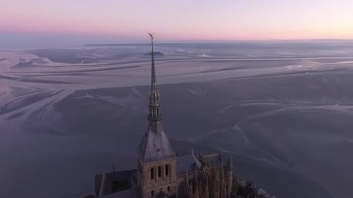 Statue of Saint Michael on spire of Mont Saint Michel abbey tower with bay in background at sunset.