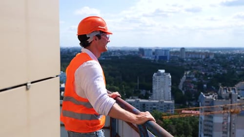 Happy Male Builder Wearing Helmet and Protective Glasses Standing on the Roof Looking at the View