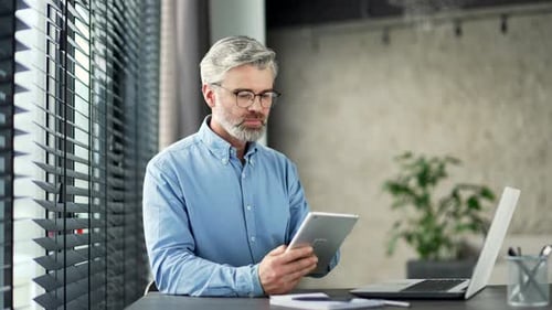 Mature gray haired bearded businessman works on a tablet while sitting at workplace in business