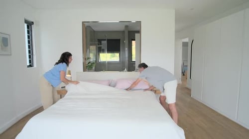 Diverse couple lifting duvet, shaking and spreading linen, smoothing pink pillows, making king bed