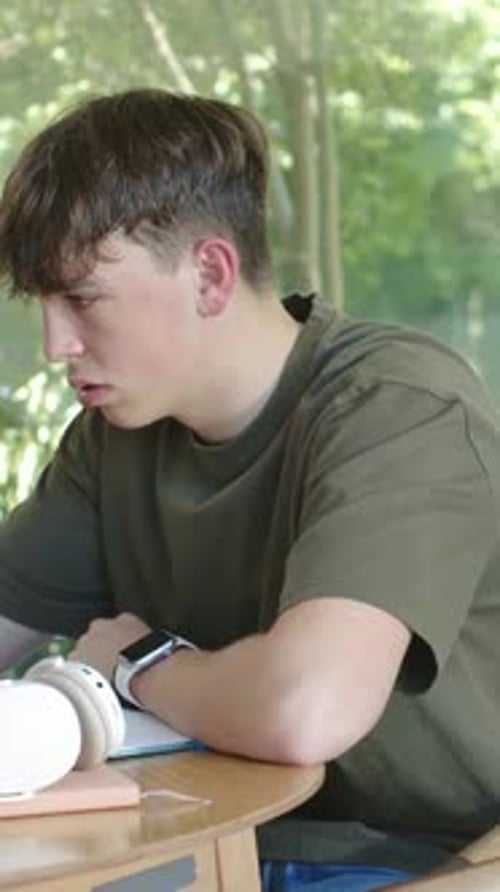 Vertical video: Leaning teen studying notebook, resting hand on temple at home window, in olive tee