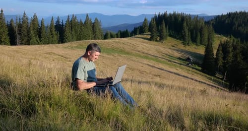 A Man Sits on a Beautiful Meadow in the Mountains Works on a Laptop