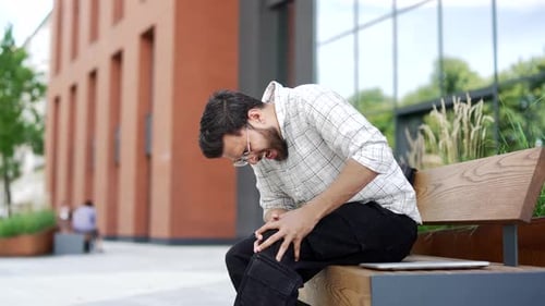 Young adult man suffers from painful severe knee joint pain sitting on a bench on street near