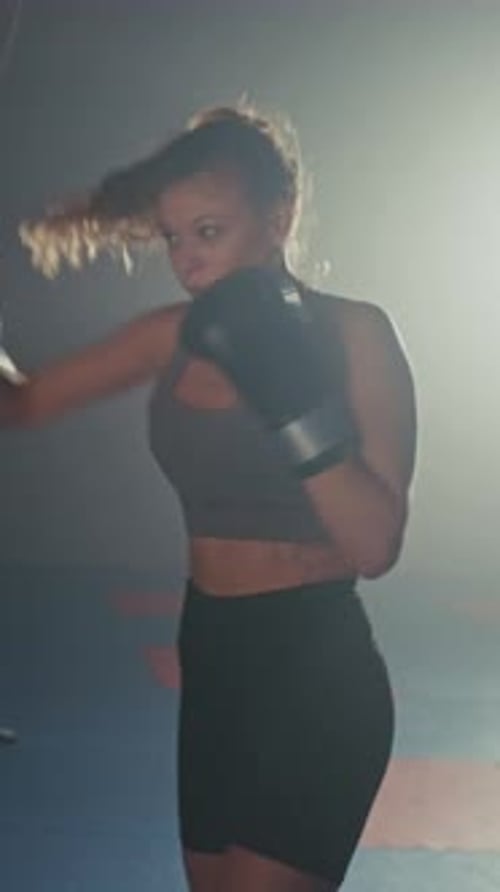 Focused Woman Trains Intensely on a Punching Bag During Evening Workout Session in a Dimly Lit Gym