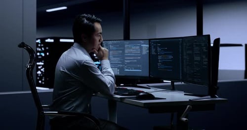 Side View Of Asian Male Programmer Thinking While Writing Code By Desktops In The Server Room
