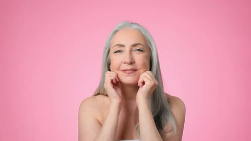Senior Woman With Gray Hair Smiling and Touching Her Face in Front of a Pink Background