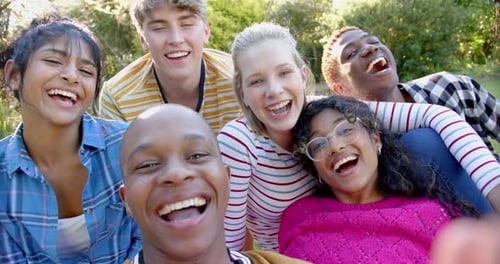 Fröhliche, vielfältige Gruppe von Freunden im Teenageralter, die auf Gras sitzen und sich im sonnigen Park umarmen, Zeitlupe