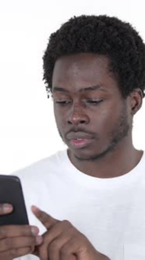 Hombre africano reaccionando ante la pérdida y usando un teléfono inteligente, fondo blanco, vídeo vertical