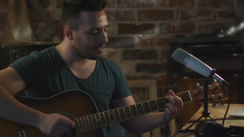 Young man is singing and playing guitar while making an audio recording at home in a garage.