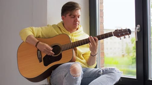 Leisure, Music And People Concept - Young Man Playing Guitar Sitting On Windowsill