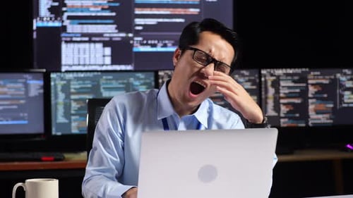 Programmer Yawning While Writing Code By A Laptop Using Multiple Monitors Showing Database