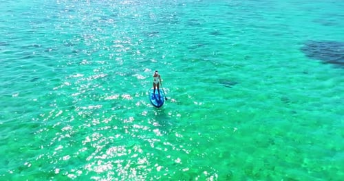 Luftaufnahme einer Frau in weißer Badesuite, die auf einem SUP-Board mit Paddel auf kristalltürkisfarbenem Wasser steht