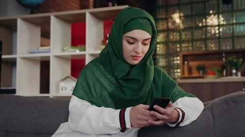 Woman in headscarf using phone on sofa indoors