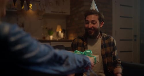 Over the Shoulder of a Happy Birthday Guy in a Festive Cap Accepting a Gift From His Girlfriend