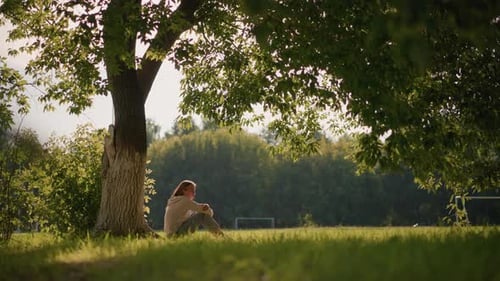 Woman Sitting By Tree in Sunlit Park Surrounded By Greenery and Gentle Breeze