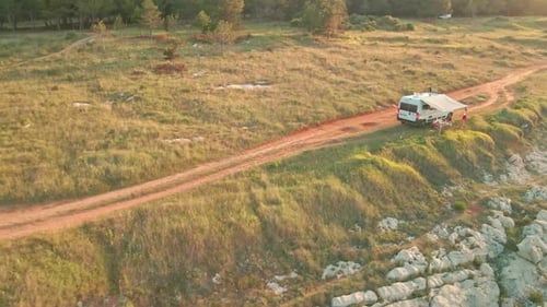 A Camper Van With Sunshade Parked Along The Dirt Road During Sunset In Green Meadows. Aerial Wide Sh