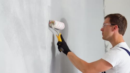 Side View of Happy Young Male Painter in Blue Overall and Gloves Painting Wall with Roller in Grey
