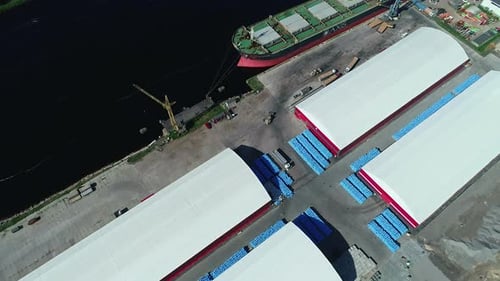 Luftaufnahme eines Frachtschiffs, das an einem sonnigen Tag am Hafen mit angrenzenden Lagerhäusern und üppigem Flussufer angedockt ist.