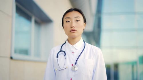 Close up portrait of Asian woman doctor looking at camera with arms crossed on background of modern