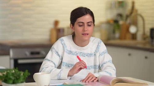 Jeune femme concentrée assise à la table de sa cuisine tout en étudiant et en prenant des notes dans son manuel
