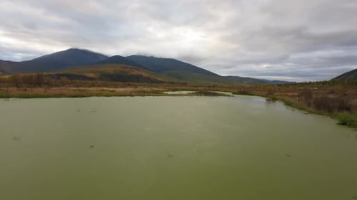 The drone soars over a calm pond reflecting the mountainous landscape, showcasing the beautiful autu