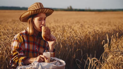 Young Girl Farmer in Sun Hat Eats Loaf of Bread Biting It Drinking Milk Sits on Wheat Field with