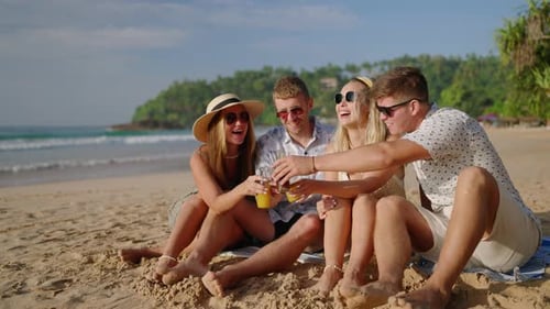 Quatre amis joyeux sont assis sur une plage de sable, boivent du kombucha et discutent lors d'une visite tropicale en riant