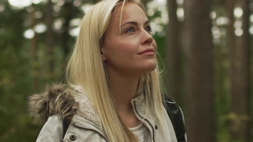 Blonde Frau im weißen Herbstmantel steht in einem Wald und schaut sich um.