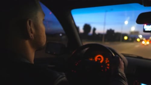 Rear View of Young Man Holds Hands on Steering Wheel and Drives a Modern Automobile at Evening Male