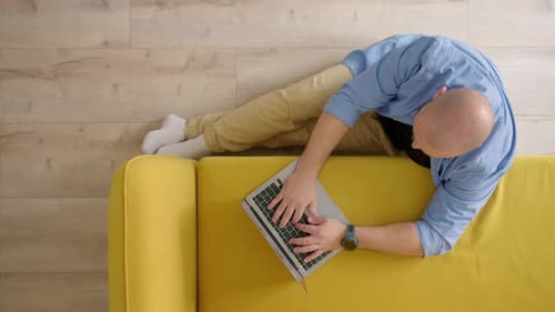 Top Down View of Sofa Next to Which Man Sits Down on Floor with Laptop and Work Unrecognizable Bald