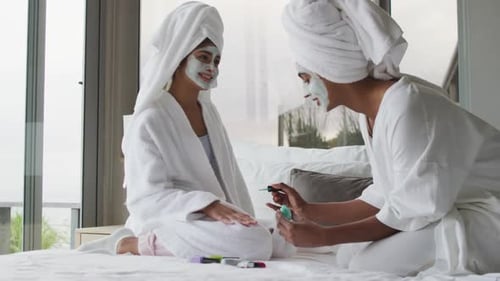 Video of happy diverse female friends moisturizing with face masks and painting nails