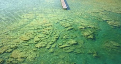 Água azul-turquesa cristalina com rochas submersas e um píer de madeira que se estende até o lago