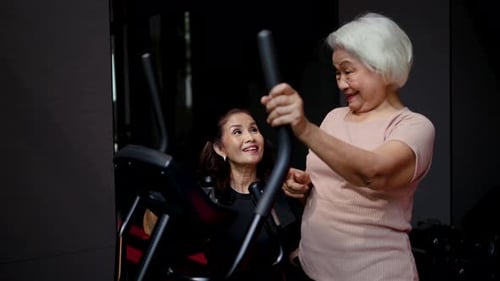 Senior Asian woman exercising on an elliptical machine with a supportive personal trainer in a gym.