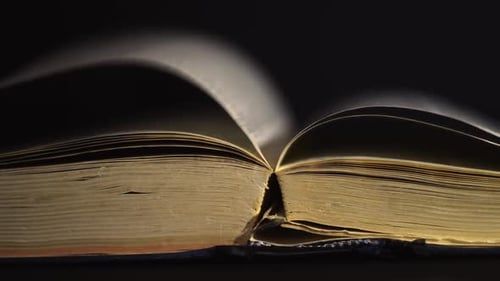 Slow motion, close up of an old, vintage, open book with pages turning on the wind, on a dark room w