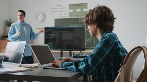 Teenage Boy Typing Code on his Laptop While Listening to IT Teacher in School