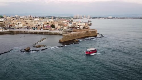 Vista aérea crepuscular de un pequeño barco rojo que cruza las antiguas murallas marinas y la arquitectura patrimonial de Acre,
