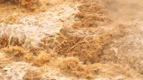 Drone aerial view of turbulent muddy floodwaters cascading over rocks.