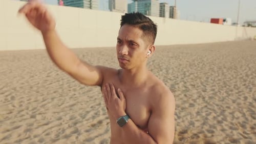 Young athletic man standing on the beach stretching his arm muscles before training