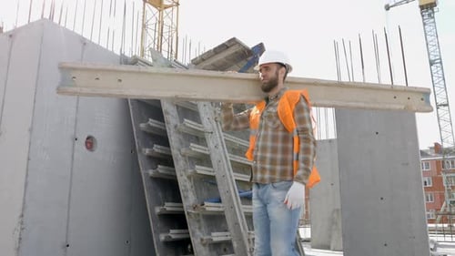 Builder man in hardhat and vest carrying timber on building site in his working day