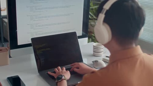 Programmer in Headphones Coding on Laptop in front of Big Monitor at Office