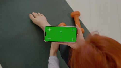 Woman takes a break on gym mat while using smartphone with green screen