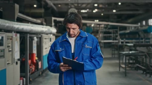 A Male Engineer in a Blue Uniform Walks Through the Factory Workshop and Checks the Operating