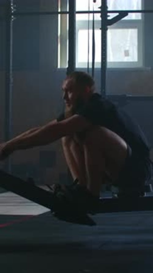 Focused Athletic Man Performing Intense Rowing Machine Workout in Dimly Lit Functional Gym