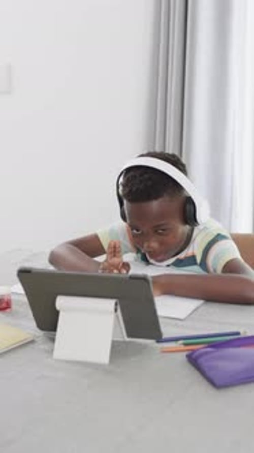 Vertical video: African American boy wearing headphones, looking at tablet and doing homework