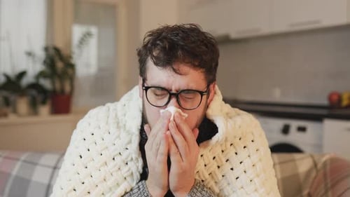 Un homme portant des lunettes fait face au rhume et à la grippe en toussant et en éternuant sous une couette