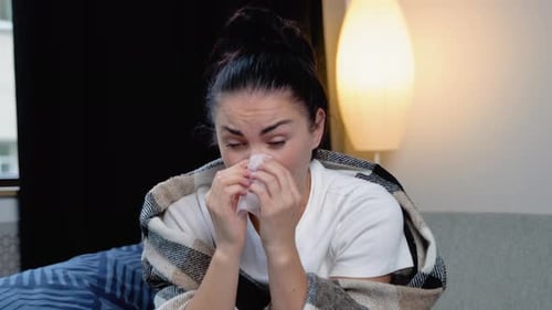 Young Woman Sits on the Couch at Home Under a Plaid Sneezing in a Napkin Feels Weak Unwell Needs