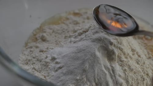 Person gießt Vanilleessenz auf einen metallischen Löffel und streut Mehl über Backmischung in einer Glasschleife