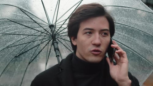 Front View of a Young Man in a Coat Walking Under a Transparent Umbrella During a Rain in the Autumn